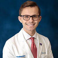 Man with short brown hair wearing a white shirt, pink tie, white coat with UT Southwestern logo, and black glasses